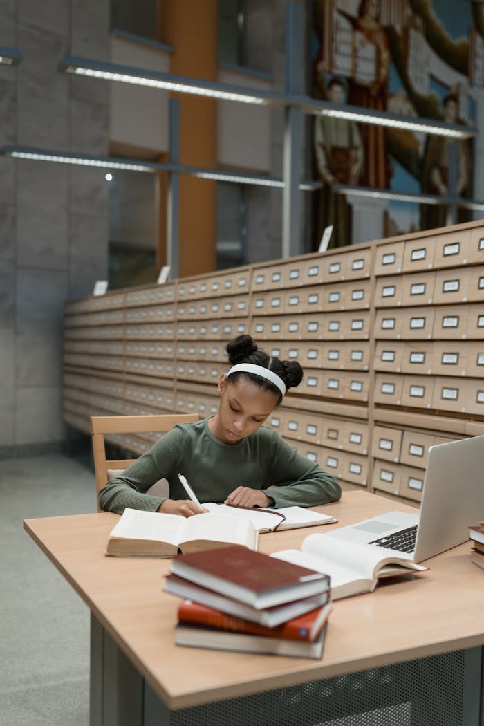 Young girl studying, writing notes in a library with books and laptop.