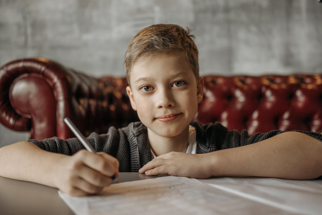 Smiling young boy writing on paper seated comfortably on a leather sofa in a cozy indoor setting.